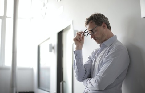 A man with his head bowed, holding glasses while leaning on a wall.