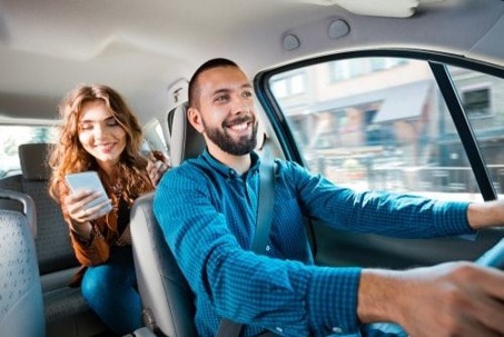 A driver smiles while giving a ride to a passenger in the backseat looking at her phone.