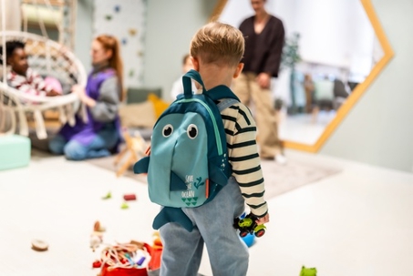 Small child with a cute save the ocean shark backpack holding a toy car, standing in a brightly lit daycare classroom surrounded by other children playing and learning