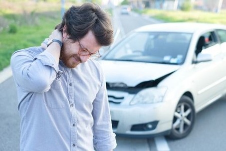 A man holds his neck in pain with a crashed car in the background.